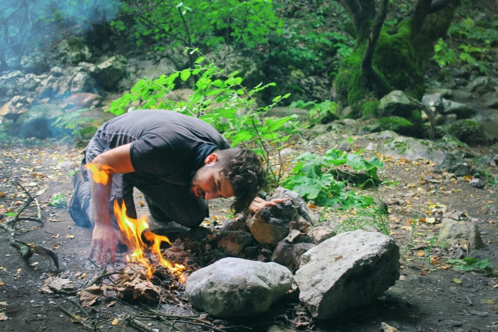 Man attempts to start a campfire surrounded by rocks in a lush green forest.