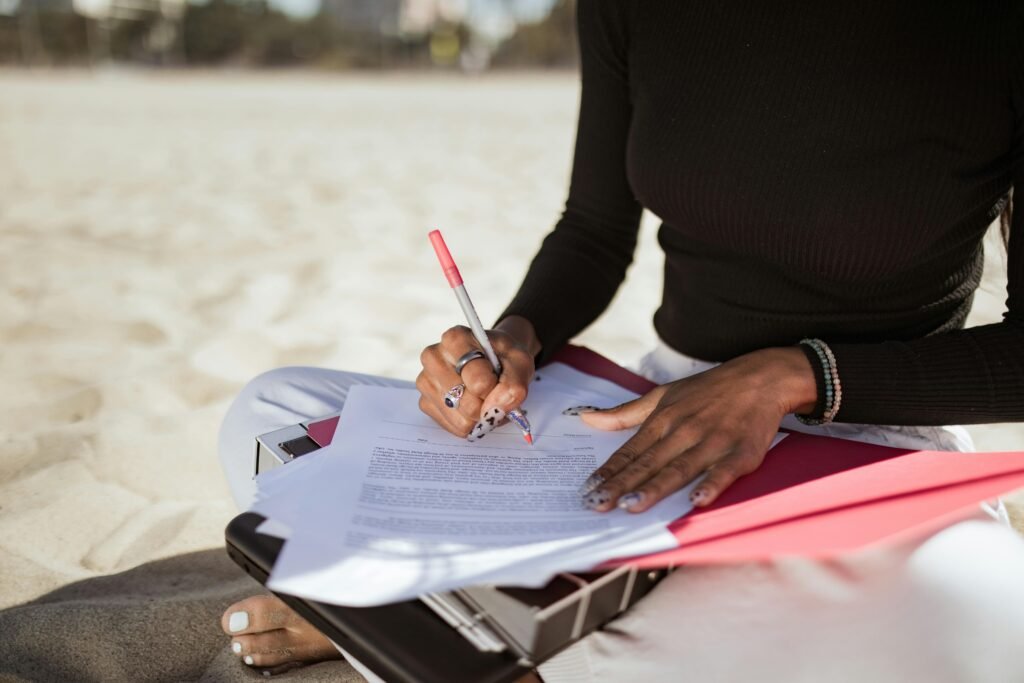 A woman writes on documents while sitting on a sandy beach, combining work and leisure.