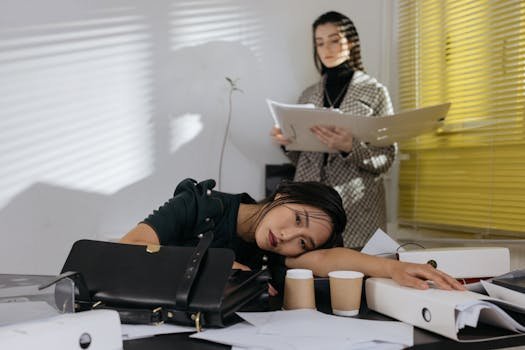 pexels photo 6538879 6538879 Exhausted employee resting on desk surrounded by documents, colleague in background.